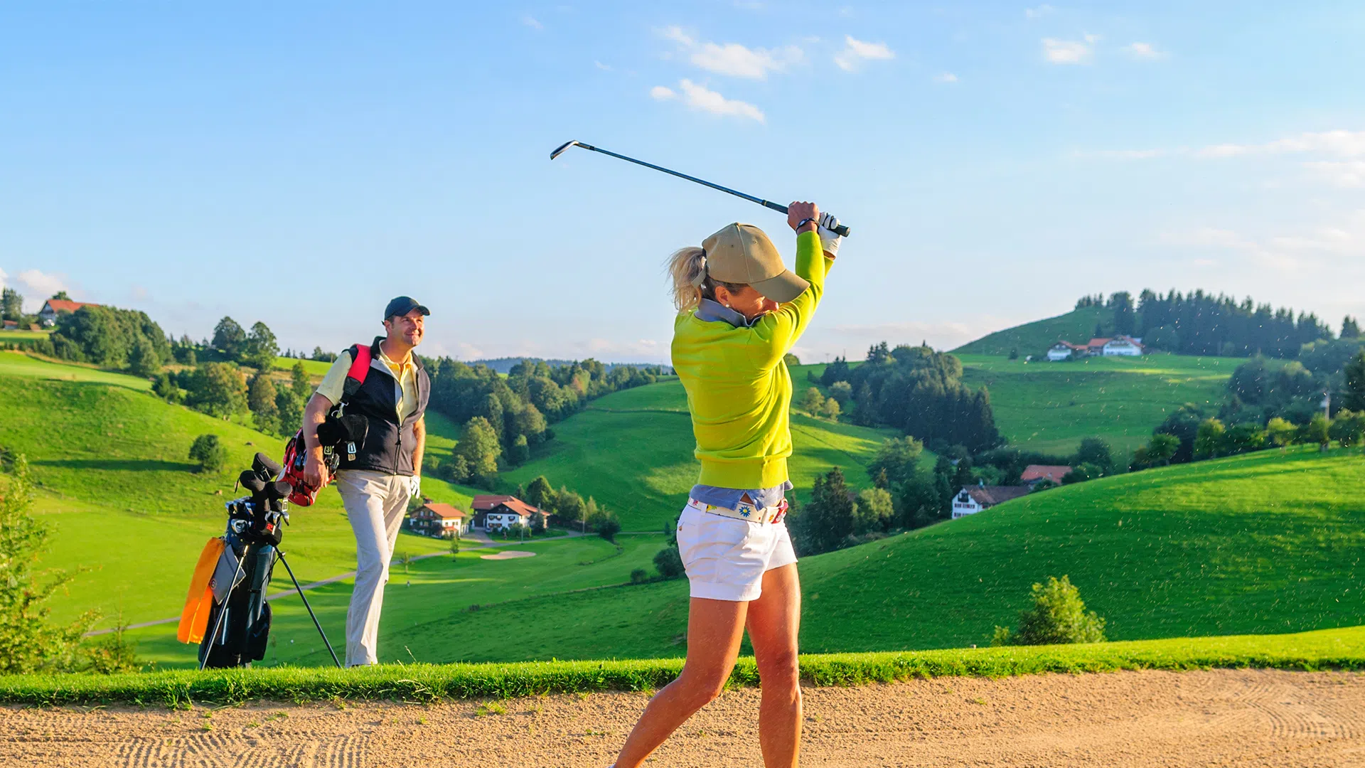 Sie sehen eine Frau und einen Mann beim Golfspielen, gerade beim Abschlag im Hintergrund mit grünen Hügeln und Sandbunker.