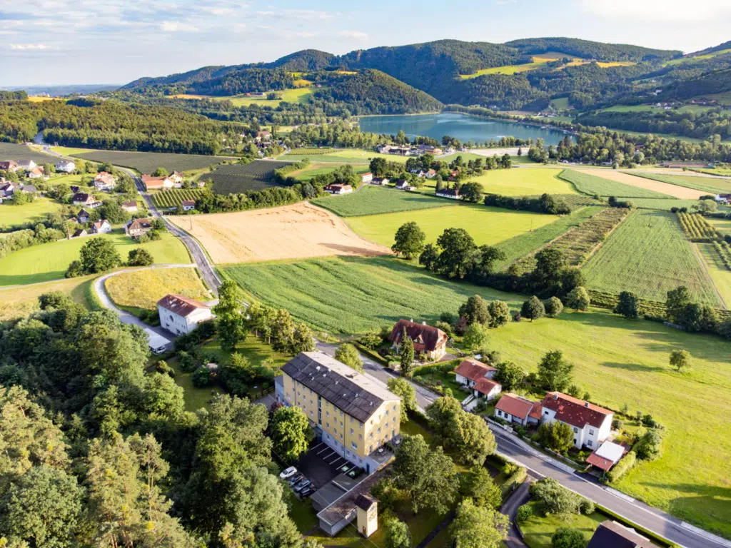 Ihr seht ein Drohnenbild vom JUFA Hotel Garni Stubenberg und den umliegenden Häusern, Wiesen und Wäldern mit blick auf den Stubenbergsee.