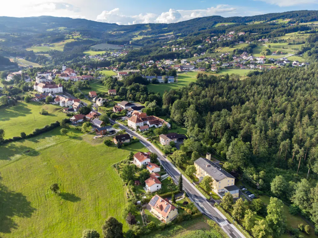 Ihr seht ein Panoramabild aus der Vogelperspektive vom Ort Stubenberg im Apfelland mit dem JUFA Hotel Garni Stubenberg, den umliegenden Wiesen und Wälder und den Hügeln der Oststeiermark.