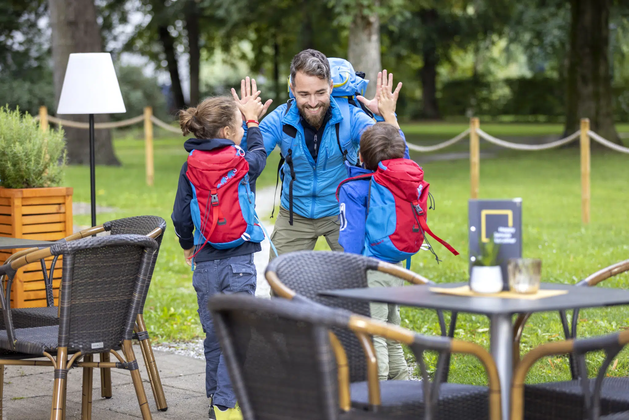 Sie sehen eine Familie, die vom Wandern zum Hotel zurückkehrt bzw. Richtung Hotel Cafe außen geht. JUFA Hotels bietet erholsamen Familienurlaub und einen unvergesslichen Winter- und Wanderurlaub.