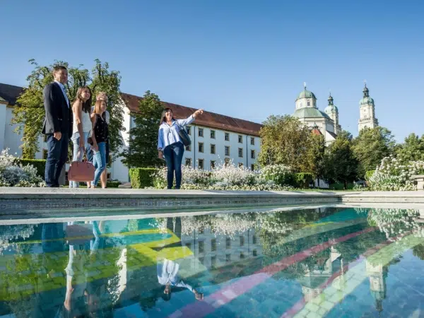 Ihr seht mehrere Personen bei der Stadtführung in Kempten. Die Familie steht gemeinsam mit der Führerin im Hofgarten Kempten.