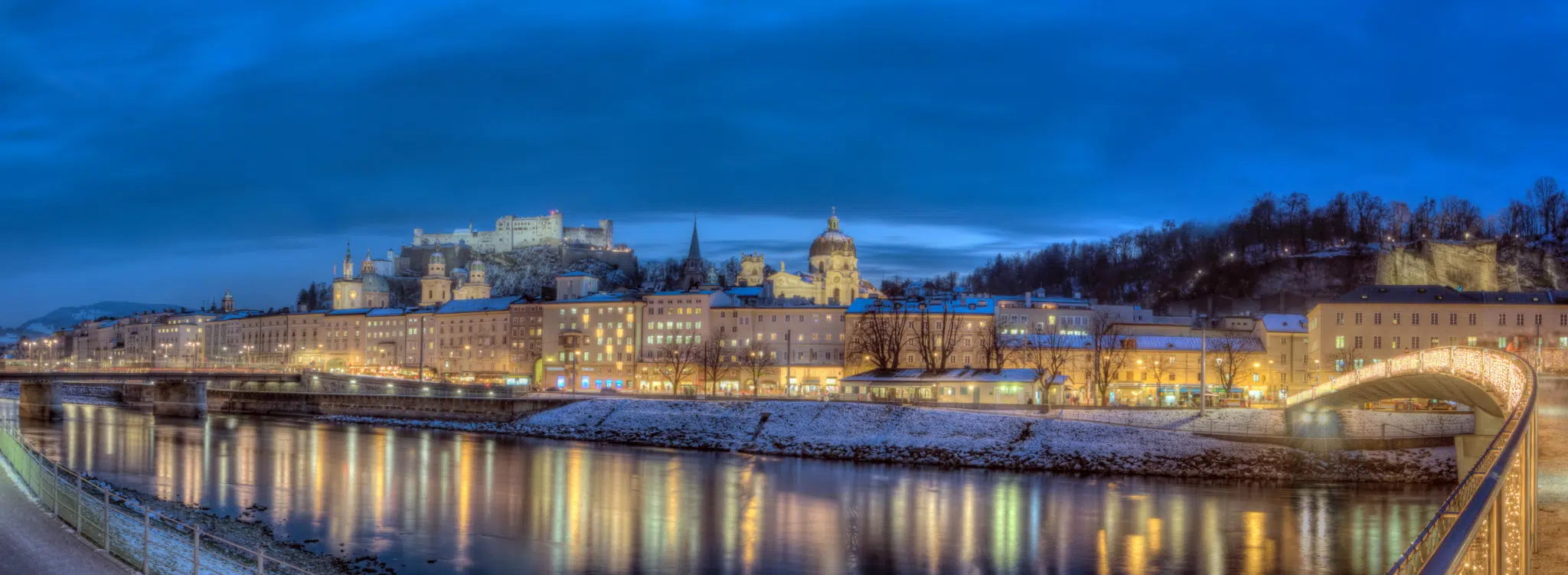 Ihr seht den Blick auf die Altstadt von Salzburg im Winter mit der Kollegienkirche, dem Salzburger Dom und der Festung Hohensalzburg sowie der Salzach im Vordergrund.