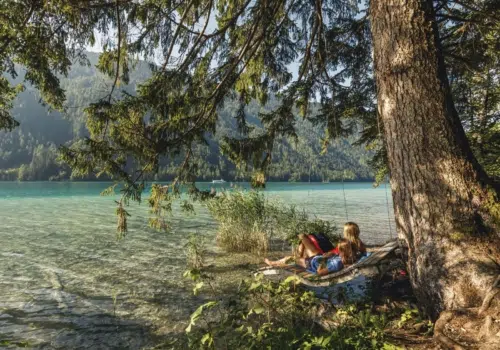 Ihr seht zwei Frauen auf einer Holzliege am Ufer des Weissensee sitzen im Sommer.