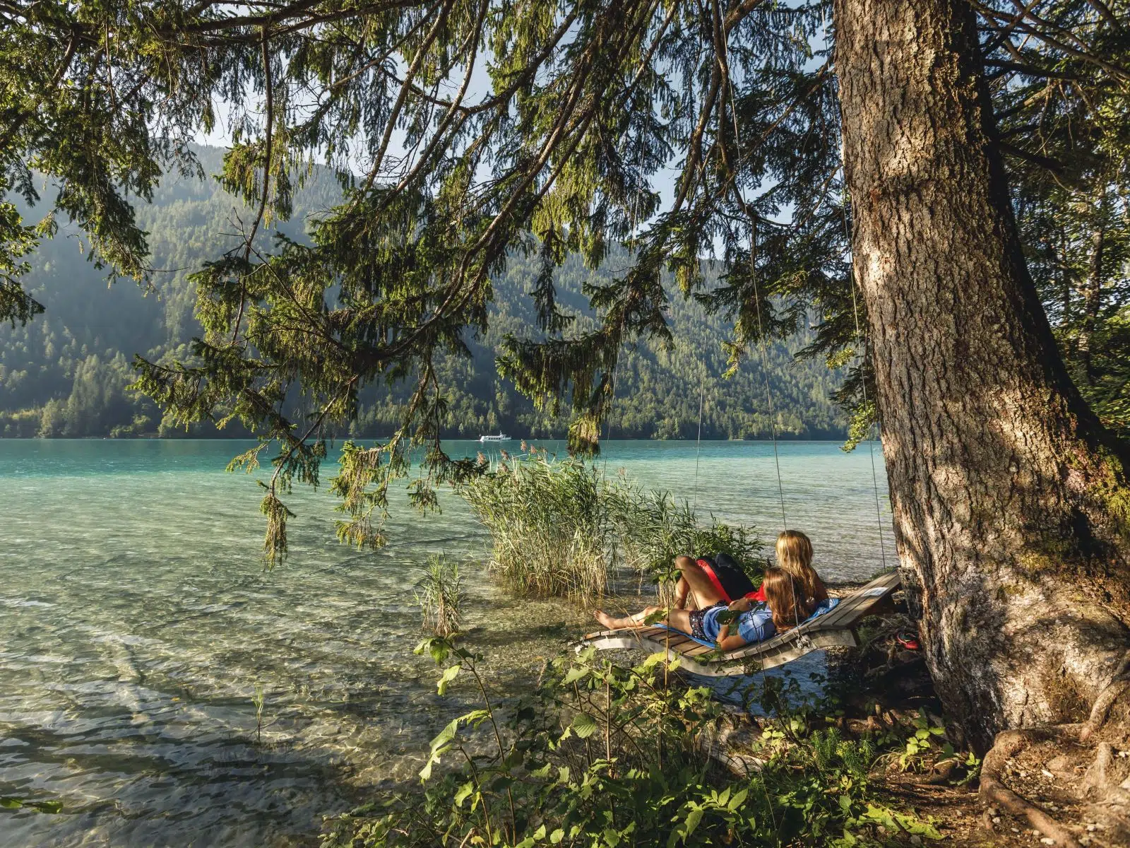 Ihr seht zwei Frauen auf einer Holzliege am Ufer des Weissensee sitzen im Sommer.