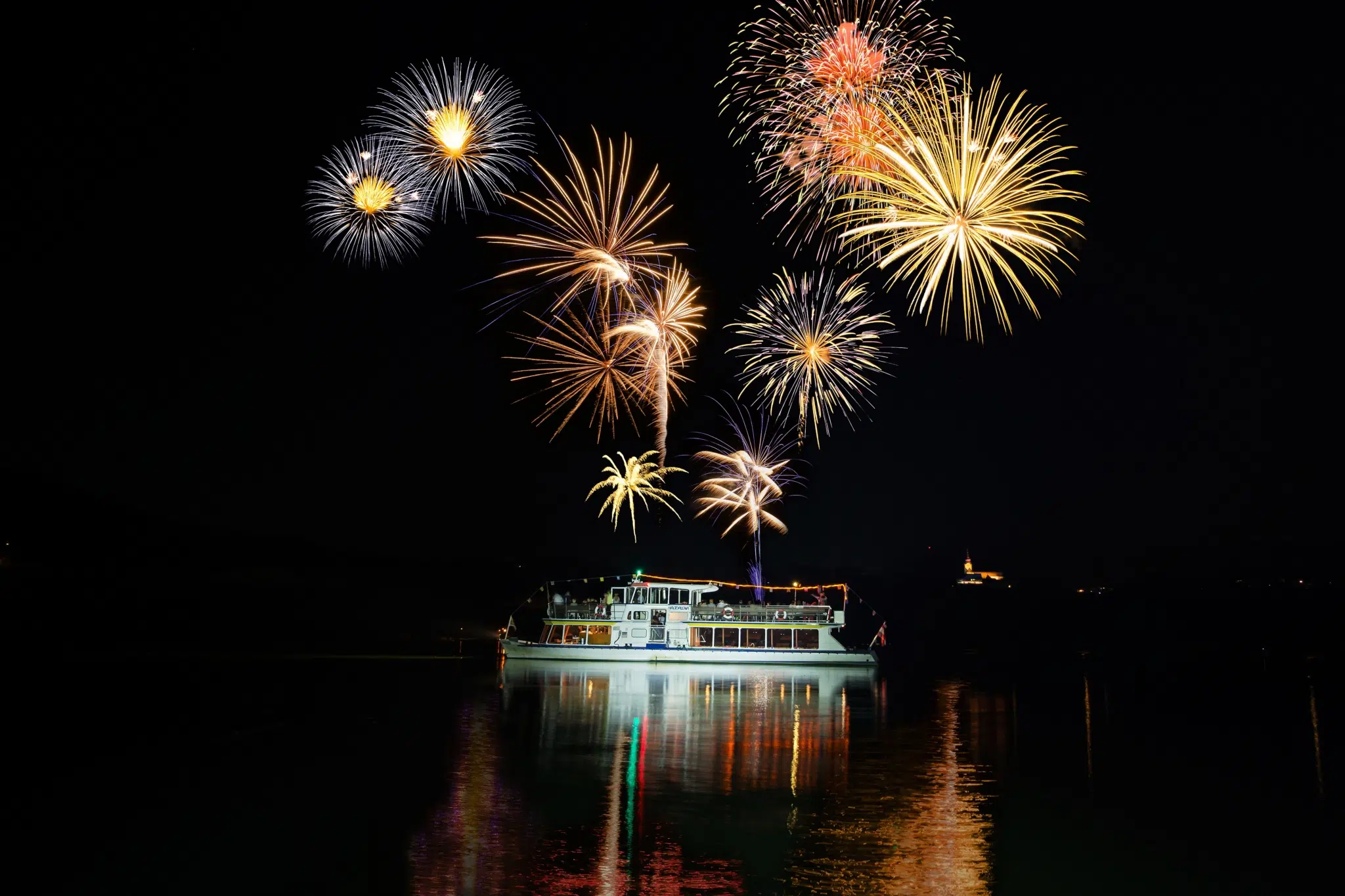 Schiff der Drauschifffahrt fährt in der Nacht auf der Drau in Südkärnten und ein Feuerwerk erleuchtet den Himmel. JUFA Hotels bieten erholsamen Familienurlaub und einen unvergesslichen Winter- und Wanderurlaub.