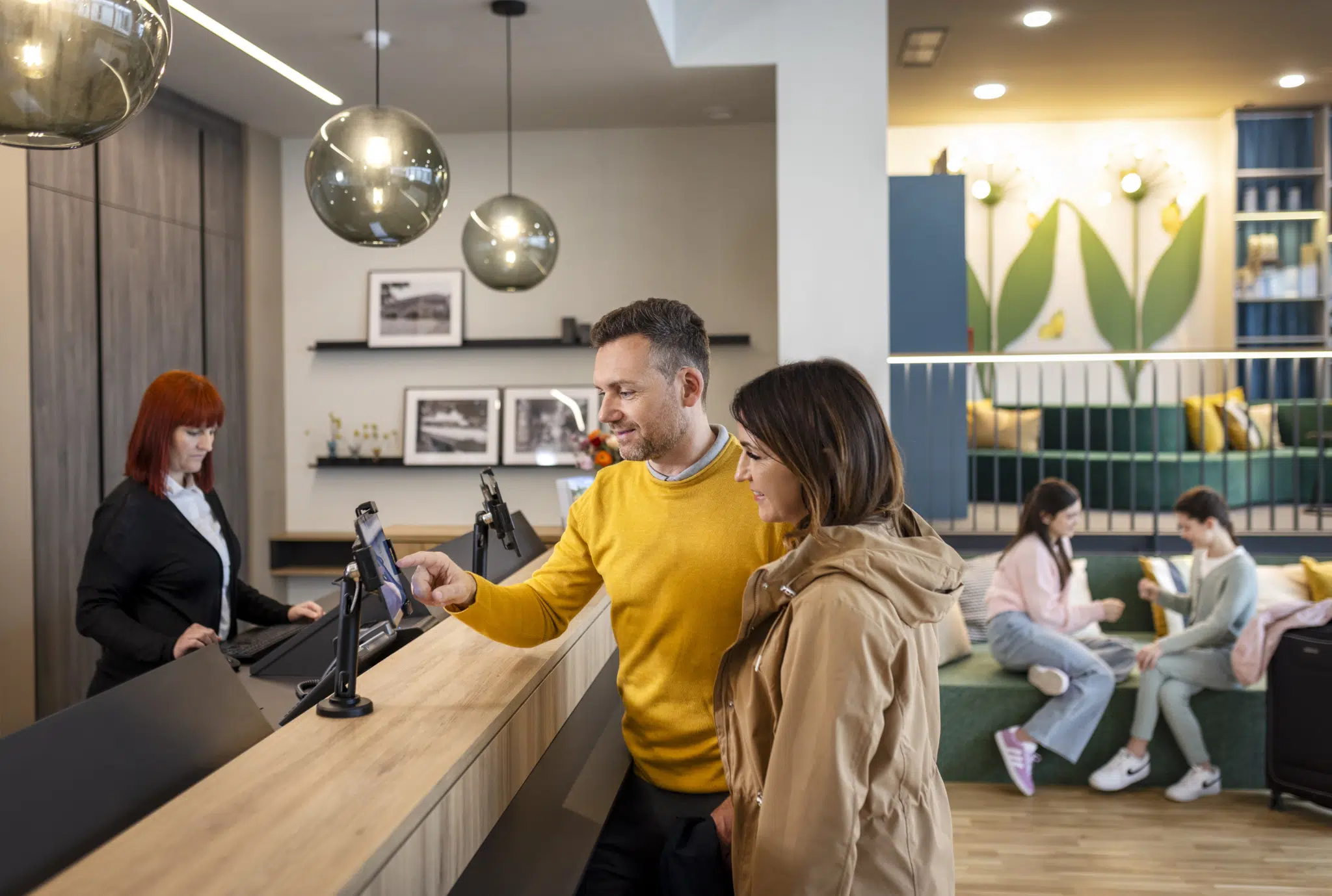 Ihr seht einen Mann und eine Frau beim Tippen am Tablet an der Rezeption im JUFA Hotel Bad Radkersburg sowie eine Mitarbeiterin und zwei Kinder im Hintergrund.