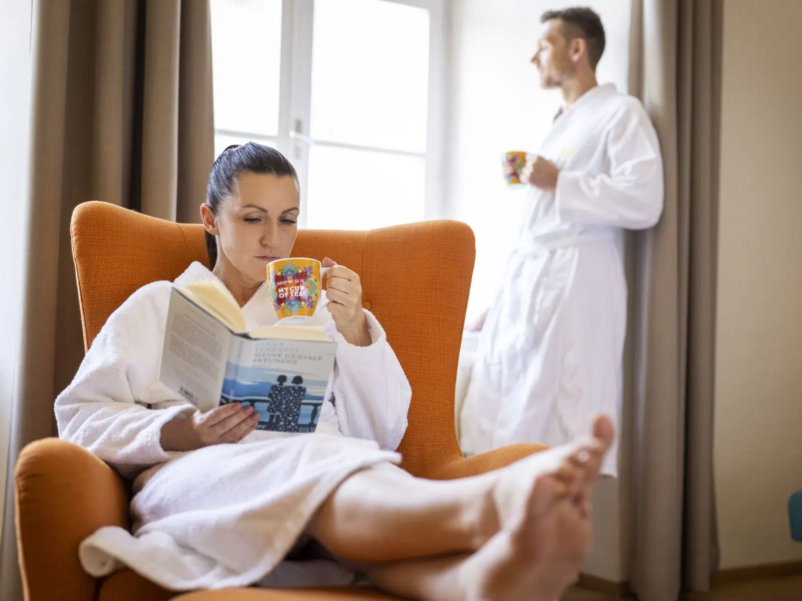 Ihr seht ein Pärchen, dass sich mit Tee und Buch am Zimmer entspannt. JUFA Hotels bietet erholsamen Familienurlaub und einen unvergesslichen Winter- und Wanderurlaub.