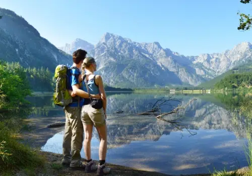 Ihr seht zwei Wanderer vor dem Bergpanorama des Almsees im Sommer.