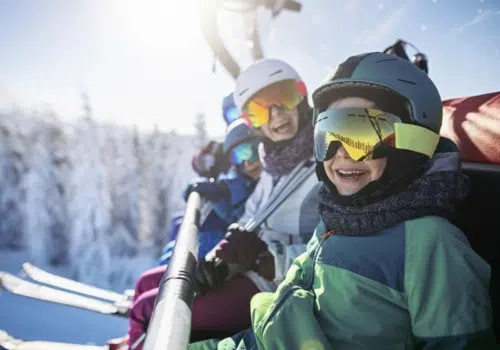 Ihr seht Kinder und Erwachsene beim Skifahren in den Alpen auf dem Sessellift.