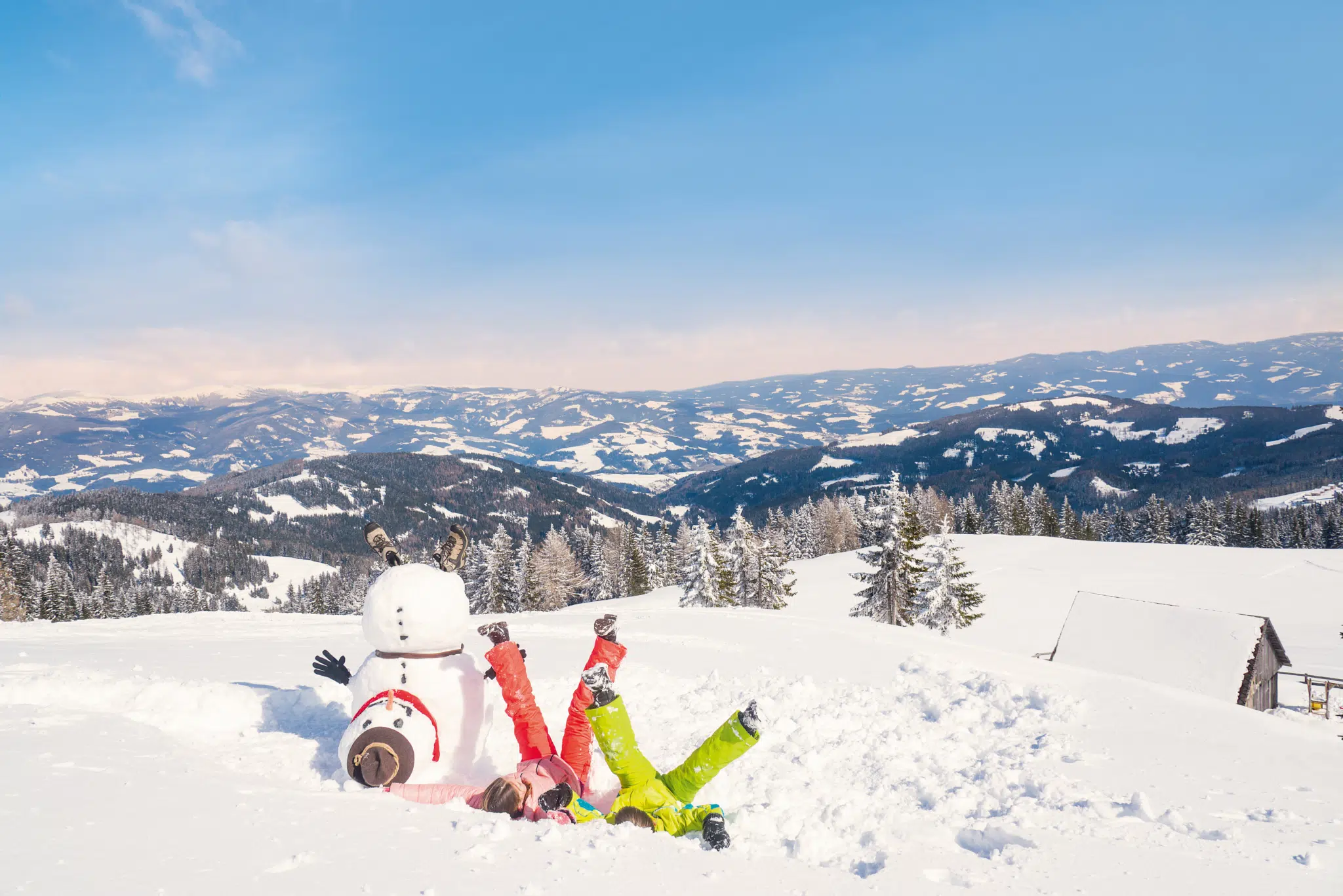 Ihr seht zwei Kinder wie sie am Rücken im Schnee liegen. Daneben steht ein Schneemann.