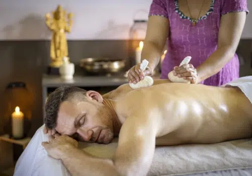 Ihr seht einen Mann, der eine Kräuterstempelmassage im Tulsi Ayurveda-Zentrum im JUFA Hotel Stift Gurk genießt. Der Ort für kinderfreundlichen und erlebnisreichen Urlaub für die ganze Familie.