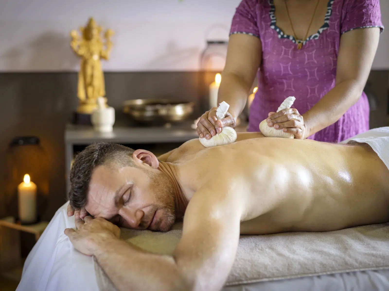 Ihr seht einen Mann, der eine Kräuterstempelmassage im Tulsi Ayurveda-Zentrum im JUFA Hotel Stift Gurk genießt. Der Ort für kinderfreundlichen und erlebnisreichen Urlaub für die ganze Familie.