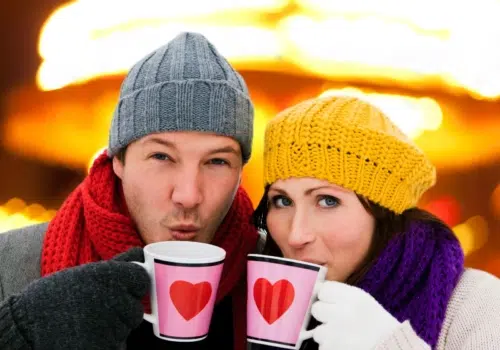 Ihr sehr ein Paar mit Hauben, das aus Herztassen Glühwein auf einem Adventmarkt trinkt. JUFA Hotels bietet erholsamen Familienurlaub und einen unvergesslichen Winterurlaub.