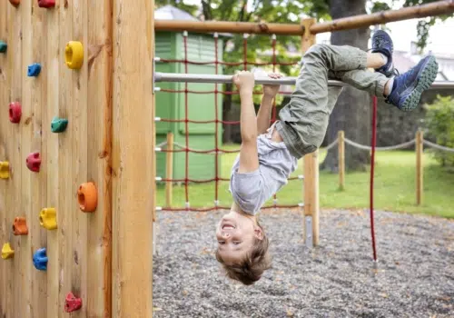 Ihr seht wie ein Kind am Spielplatz auf der Kletterstange kopfüber hängt und lacht.