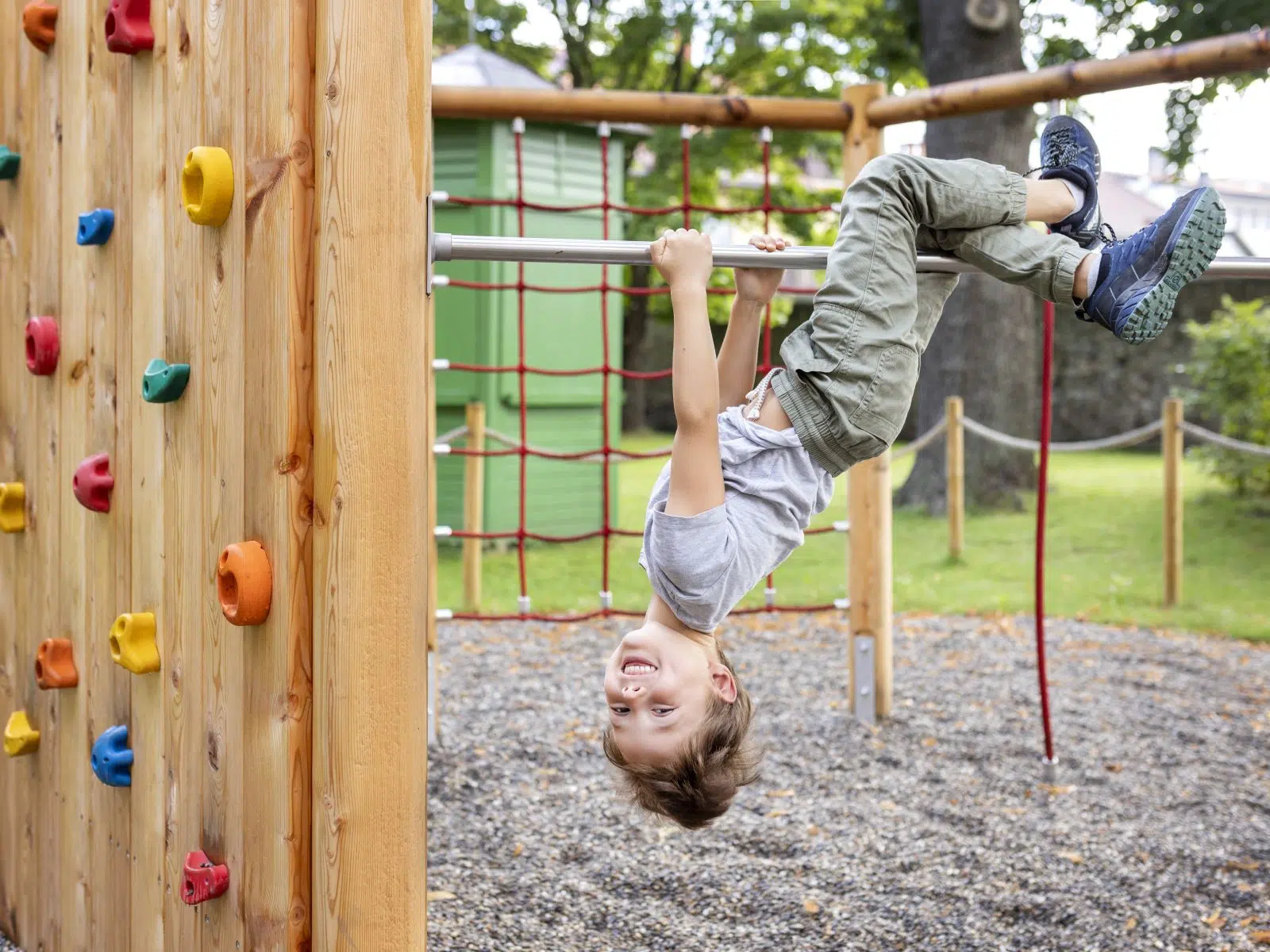 Ihr seht wie ein Kind am Spielplatz auf der Kletterstange kopfüber hängt und lacht.