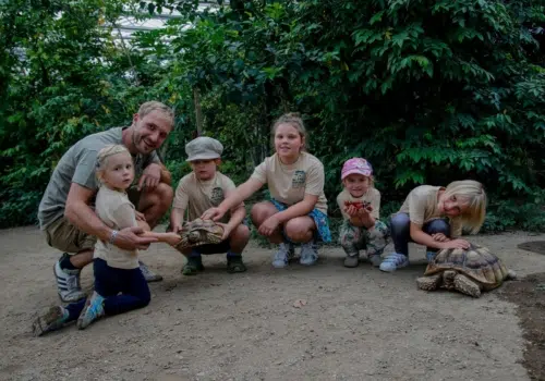 Ihr seht eine Gruppenaufnahme im Tropenhaus am Rennsteig mit Kindern, einem Gruppenführer und einer Schildkröte. JUFA Hotels bietet kinderfreundlichen und erlebnisreichen Urlaub für die ganze Familie.