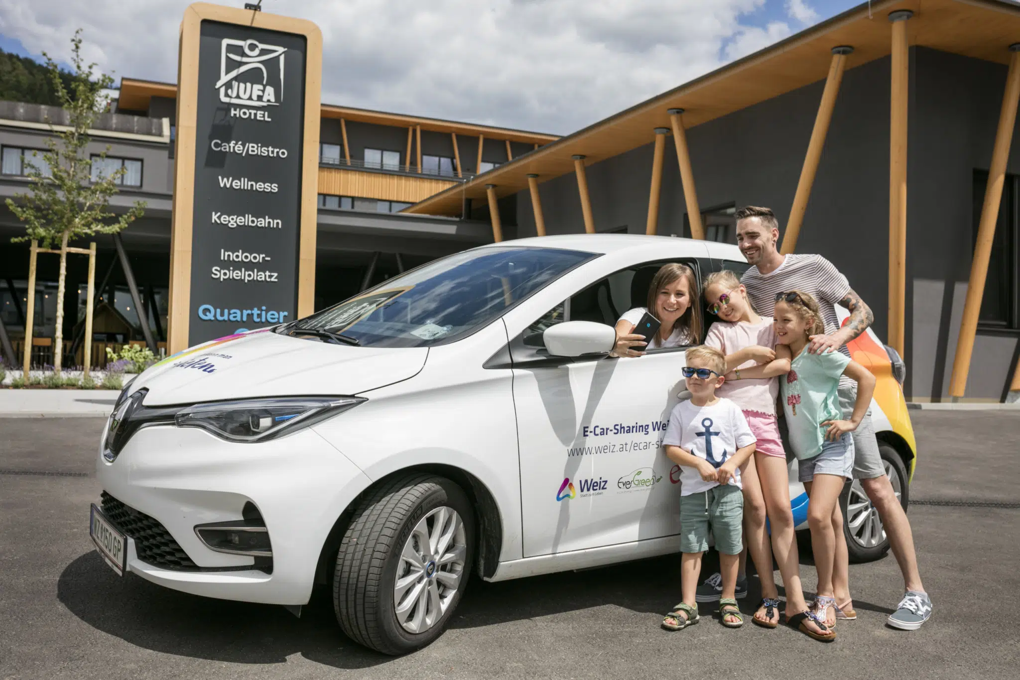 Ihr seht eine Familie mit Elektroauto vor dem JUFA Hotel. Der Ort für erholsamen Familienurlaub und einen unvergesslichen Winter- und Wanderurlaub.