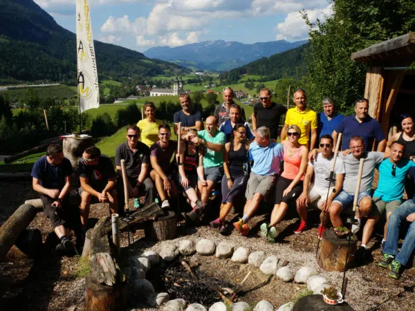 Eine Gruppe Erwachsener sitzt in Pyhrn-Priel im Wald im Outdoor Camp für ein Gruppenfoto zusammen. JUFA Hotels bietet den Ort für erfolgreiche und kreative Seminare in abwechslungsreichen Regionen.