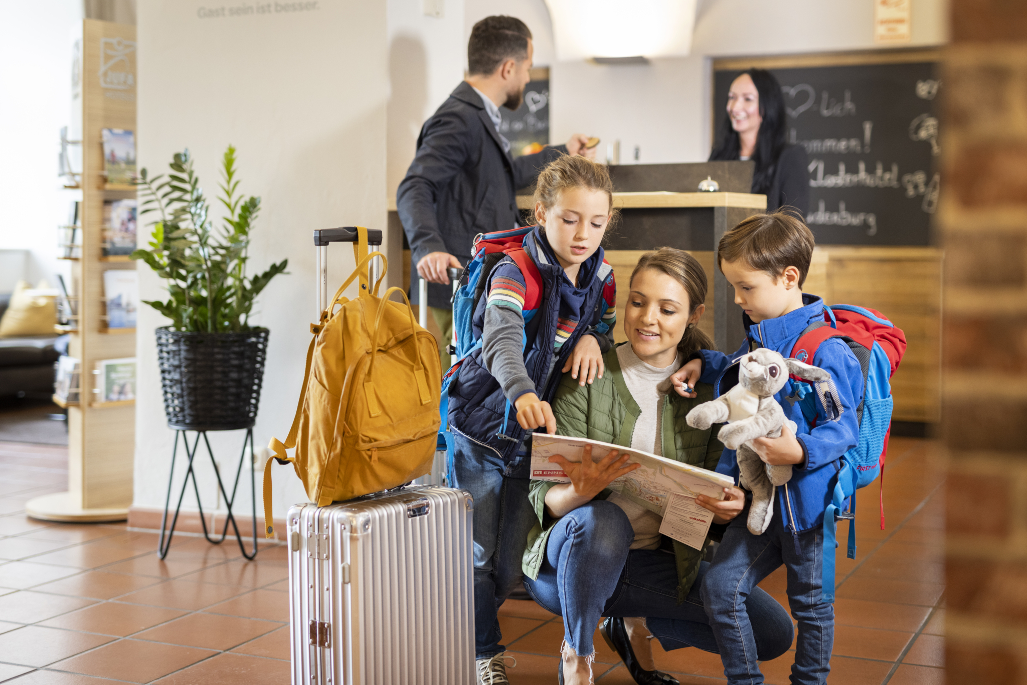 Ihr seht eine Mutter mit ihren Kindern bei der Ankunft im Hotel. Sie sehen sich gemeinsam eine Karte an während der Vater den Checkin regelt. JUFA Hotels bietet erholsamen Familienurlaub und einen unvergesslichen Winter- und Wanderurlaub.