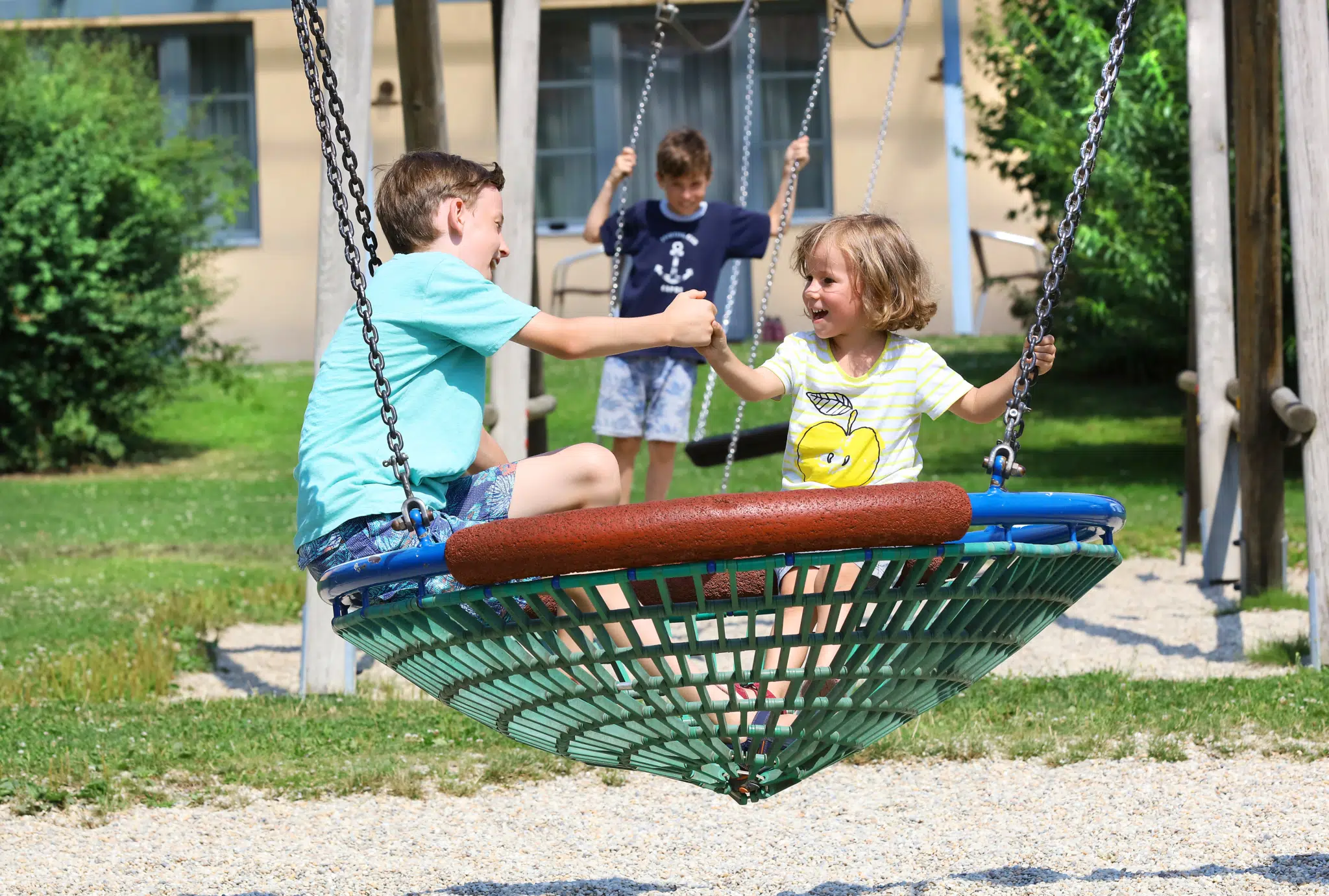 Ihr seht Kinder auf einer Korbschaukel am Spielplatz des JUFA Hotels Pöllau - Bio-Landerlebnis. Der Ort für erholsamen Familienurlaub und einen unvergesslichen Winter- und Wanderurlaub.