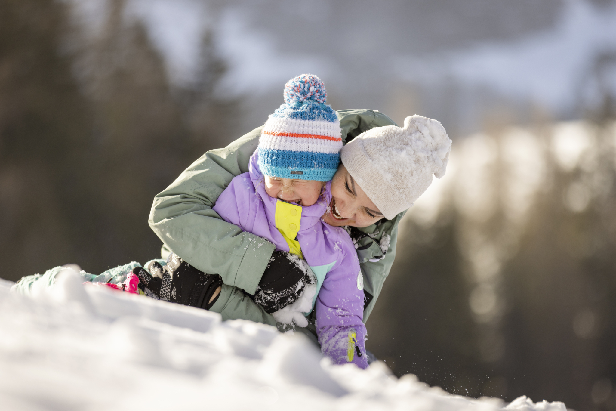 Ihr seht eine Frau, die lachend ein Mädchen umarmt, im Schnee sitzend.