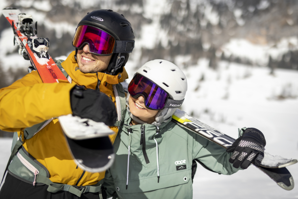 Ihr seht einen Mann und eine Frau in Skikleidung mit Helm und Skibrille, die Ski über der Schulter tragen, in verschneiter Winterlandschaft.