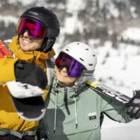 Ihr seht einen Mann und eine Frau in Skikleidung mit Helm und Skibrille, die Ski über der Schulter tragen, in verschneiter Winterlandschaft.