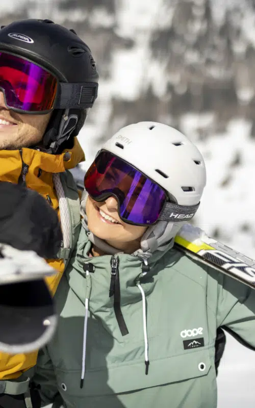 Ihr seht einen Mann und eine Frau in Skikleidung mit Helm und Skibrille, die Ski über der Schulter tragen, in verschneiter Winterlandschaft.