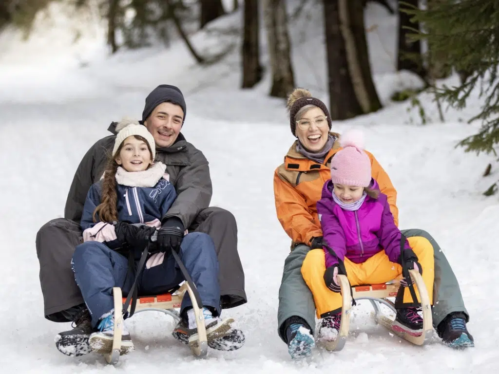 Ihr seht eine Familie beim Schlittenfahren durch den Wald. Das JUFA Hotel Laterns ist ein idealer Ort für einen unvergesslichen Winter- und Familienurlaub und erholsame Tage für die ganze Familie.