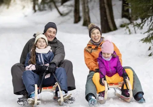 Ihr seht eine Familie beim Schlittenfahren durch den Wald. Das JUFA Hotel Laterns ist ein idealer Ort für einen unvergesslichen Winter- und Familienurlaub und erholsame Tage für die ganze Familie.