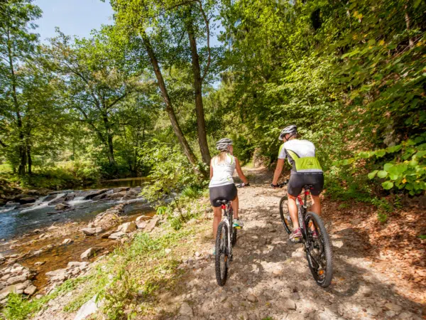 Ihr seht zwei Personen beim Radfahren auf einem Waldweg neben einem Fluss.