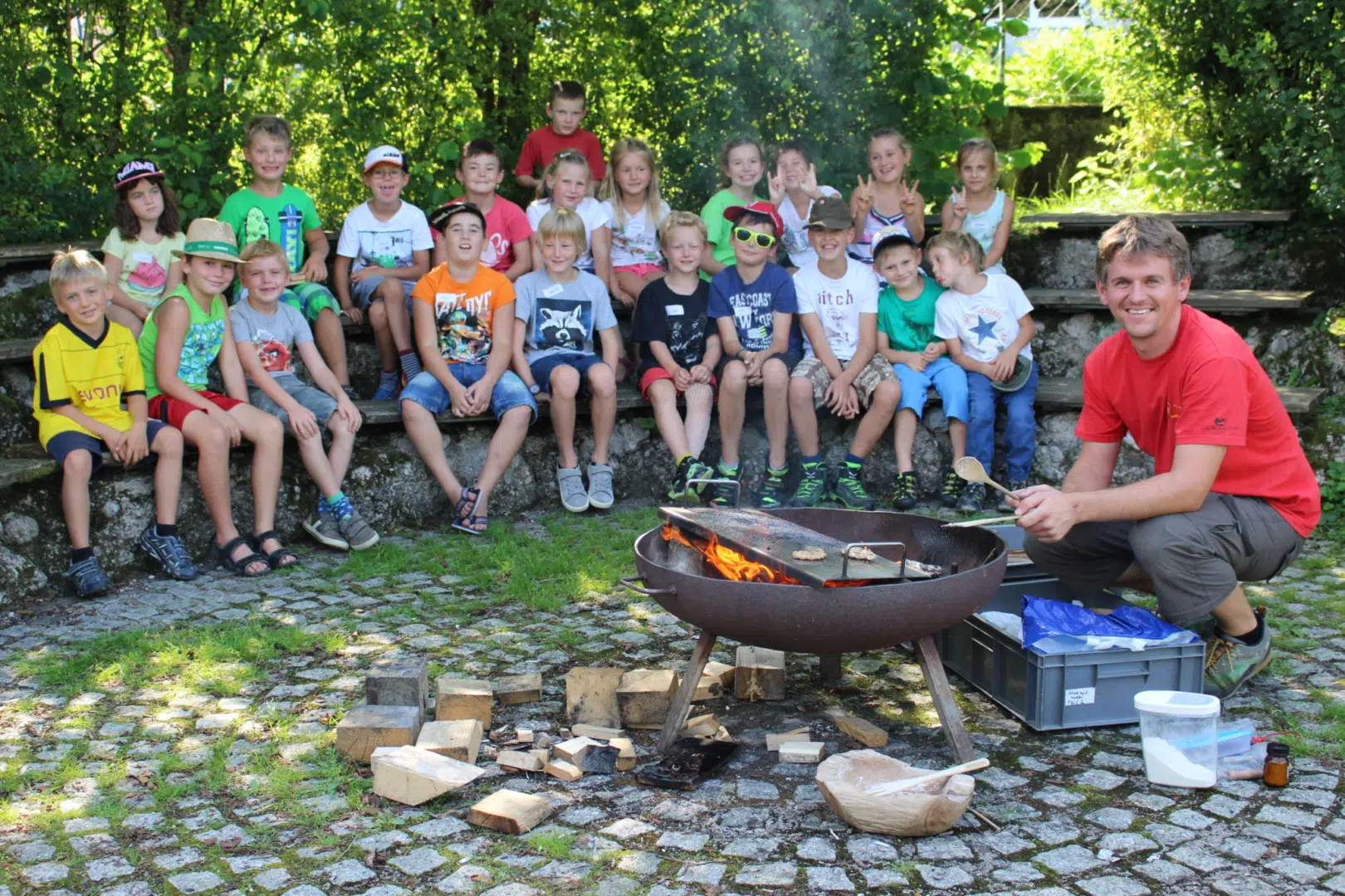 Sie sehen eine Gruppe Kinder mit Betreuer von abenteuer management beim Grillen. JUFA Hotels bietet erlebnisreiche und kreative Schulprojektwochen in abwechslungsreichen Regionen.