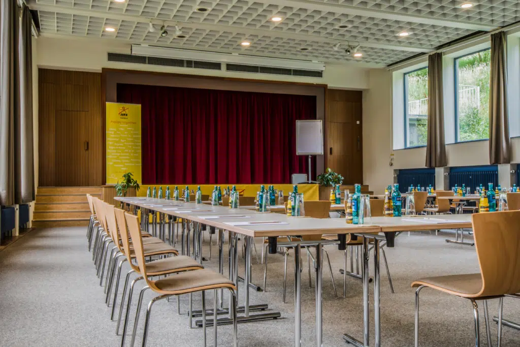 Ihr seht einen gut ausgestatteter Seminarraum mit Bühne und Seminargetränken im JUFA Hotel Königswinter/Bonn. Der Ort für erlebnisreichen Städtetrip für die ganze Familie und der ideale Platz für Ihr Seminar.