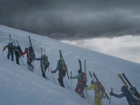 Ihr seht ein Standbild aus dem Film "Todesfall Haute Route": Eine Gruppe von Skitourengehern stapft einen schneebedeckten Hang hinauf, im Hintergrund dunkle Wolken.