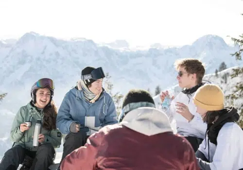 Ihr seht eine Gruppe von Snowboarder*innen, die lachend am Kasberg in Oberösterreich Pause im Schnee machen.