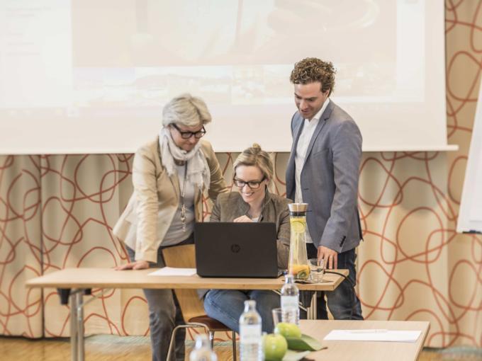 Zwei Seminarteilnehmer und die Seminarleiterin versammeln sich um einen Laptop im JUFA Hotel Wien City. Der Ort für erlebnisreichen Städtetrip für die ganze Familie und der ideale Platz für Ihr Seminar.
